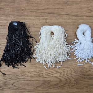 Puede incluir: Tres haces de cuerda, uno negro, uno blanco y uno crema, están dispuestos sobre una superficie de madera. Las cuerdas están atadas juntas en la parte superior y se abren en abanico en la parte inferior.