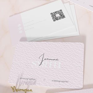 May include: A stack of white business cards with a light pink side accent, a QR code, and contact icons. A textured light pink card in the foreground displays the name Joanne Smith, the titles photographer and videographer, and a gold script nameplate.