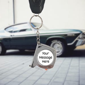 Può includere: Un portachiavi a metro a nastro argentato con un cerchio bianco vuoto con la scritta "Your Message Here". Il portachiavi è appeso a una chiave dell'auto. Un'auto classica verde scuro è sullo sfondo.