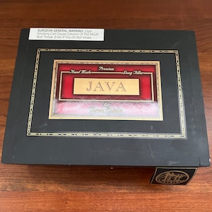 May include: A black wooden box with a gold trim and a red label that says "JAVA by Dram Estate" with the words "Hand Made Premium Long Filler" above the name. The box has a Surgeon General's Warning label on the top.