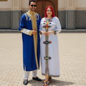 May include: Two people wearing traditional Moroccan attire. The man is in a blue robe with gold trim, white pants, and black shoes. The woman is in a white dress with colorful embroidery and patterned shoes. They are standing in front of a building.