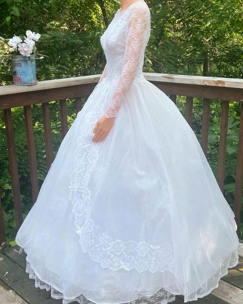May include: A white wedding dress with a full skirt and long lace sleeves. The bodice features delicate lace detailing, and the skirt has a layered, flowing design. The dress is suitable for a wedding or special occasion.