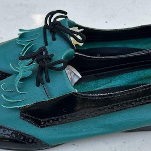 May include: A pair of teal and black lace-up shoes featuring a fringe tongue detail, a rounded toe, and black patent leather accents with decorative brogue-style perforations along the edges. The shoes include thin black laces and a flat sole.
