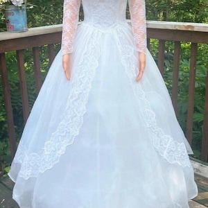 May include: A white wedding dress with a lace bodice and long lace sleeves. The dress features a full skirt with lace trim and a delicate lace neckline. The dress is made of a sheer fabric and appears to be vintage.