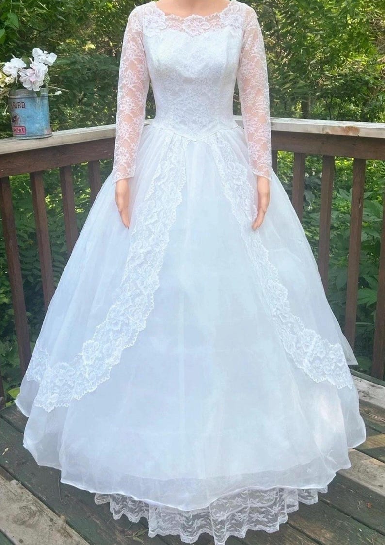 May include: A white wedding dress with a lace bodice and long lace sleeves. The dress features a full skirt with lace trim and a delicate lace neckline. The dress is made of a sheer fabric and appears to be vintage.
