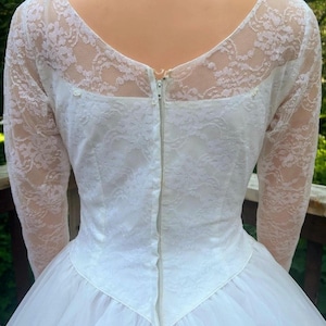 May include: A vintage white wedding dress with a lace bodice and long sleeves. The back features a zipper closure and a tulle skirt. The dress has a classic, elegant design.