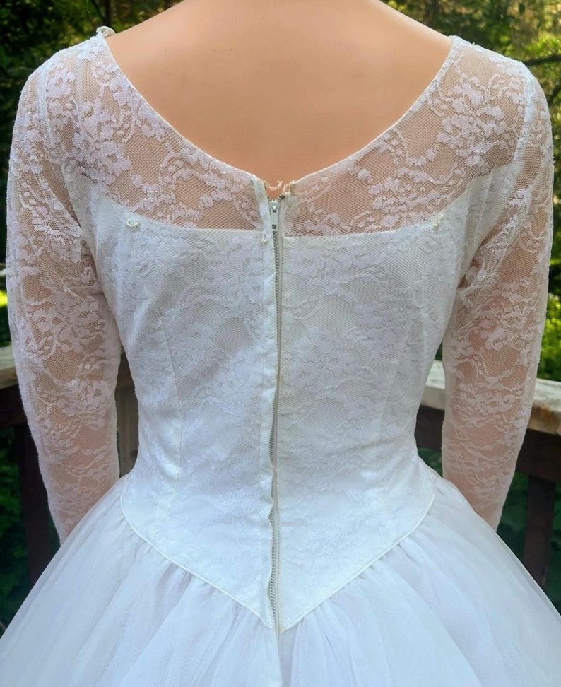 May include: A vintage white wedding dress with a lace bodice and long sleeves. The back features a zipper closure and a tulle skirt. The dress has a classic, elegant design.