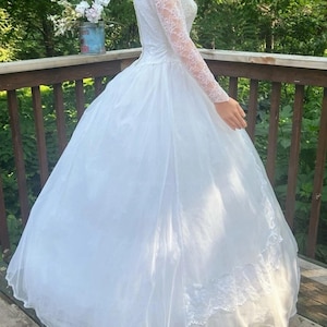 May include: A white wedding dress with a full skirt and long lace sleeves. The dress has a fitted bodice and a flowing skirt. The sleeves are made of delicate lace. The dress is made of a lightweight fabric and appears to be vintage.