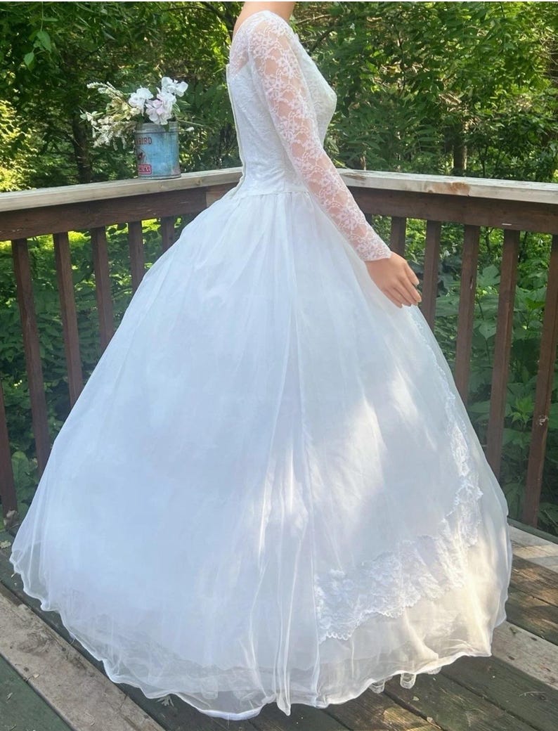 May include: A white wedding dress with a full skirt and long lace sleeves. The dress has a fitted bodice and a flowing skirt. The sleeves are made of delicate lace. The dress is made of a lightweight fabric and appears to be vintage.