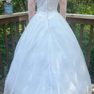 May include: A white wedding dress with a full skirt and long lace sleeves. The back of the dress features a zipper closure and a V-neckline. The dress is made of a lightweight fabric, and the skirt has multiple layers.