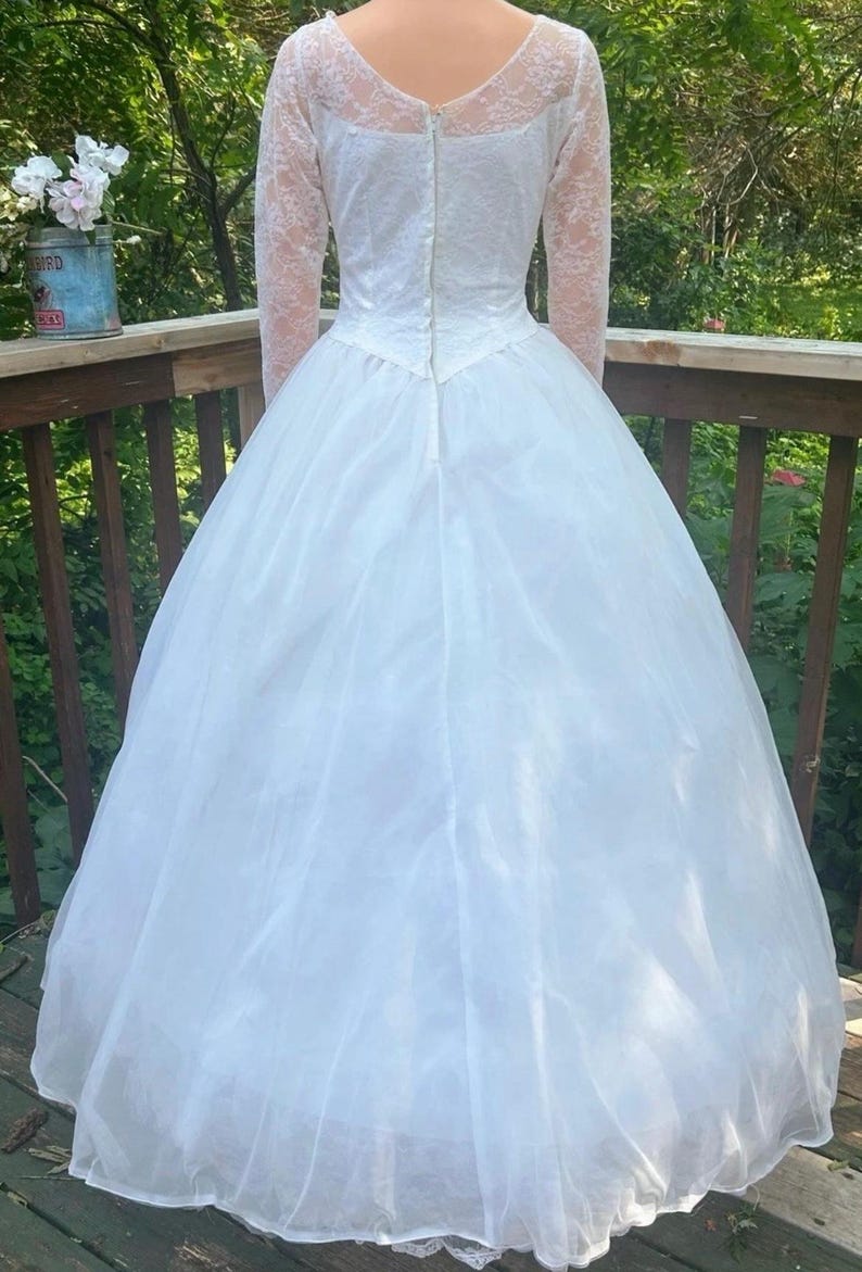 May include: A white wedding dress with a full skirt and long lace sleeves. The back of the dress features a zipper closure and a V-neckline. The dress is made of a lightweight fabric, and the skirt has multiple layers.