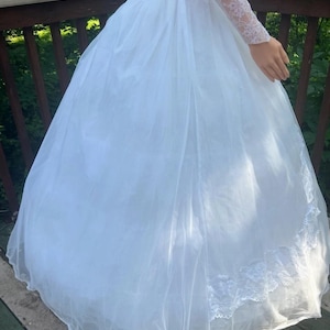 May include: A white wedding dress with a full skirt and lace trim. The dress features long, lace sleeves and a high neckline. The skirt is made of layers of sheer fabric, creating a voluminous silhouette. The dress is designed for a formal occasion.
