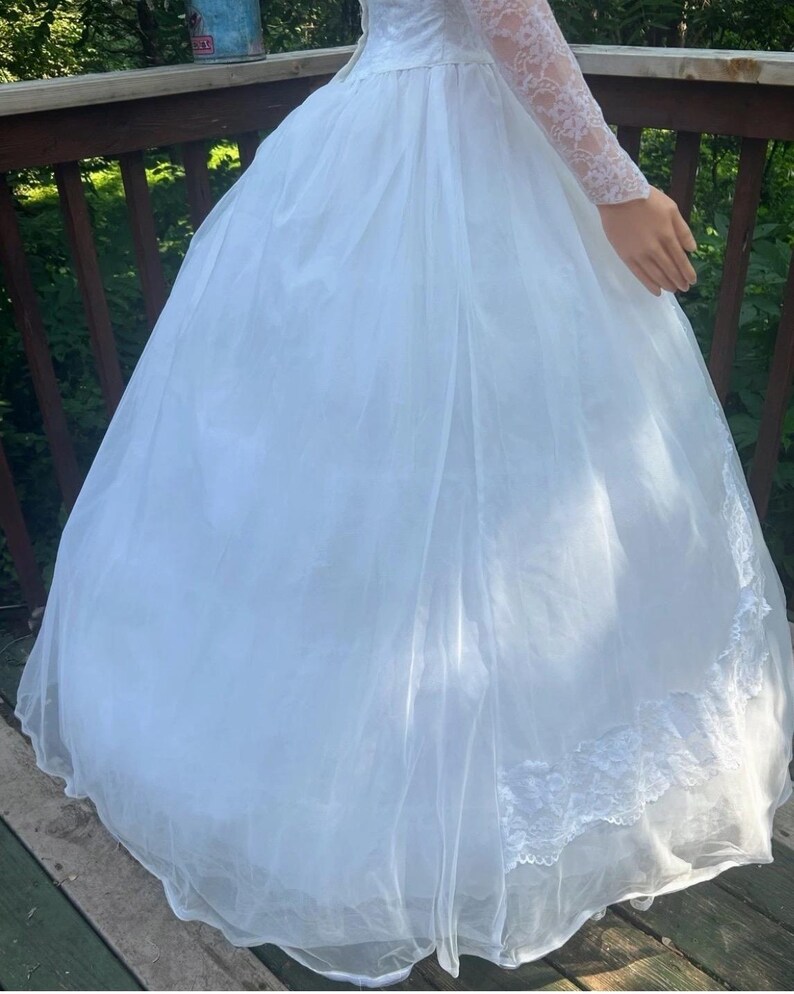 May include: A white wedding dress with a full skirt and lace trim. The dress features long, lace sleeves and a high neckline. The skirt is made of layers of sheer fabric, creating a voluminous silhouette. The dress is designed for a formal occasion.