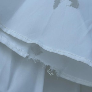 May include: Close-up of white fabric with a torn section and a decorative cut-out. The fabric appears to be a light, possibly cotton or linen material, with visible stitching along the edges. The torn area reveals the fabric's internal structure.