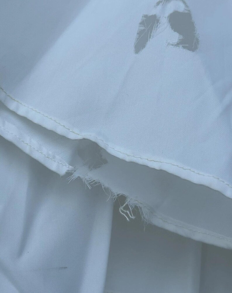 May include: Close-up of white fabric with a torn section and a decorative cut-out. The fabric appears to be a light, possibly cotton or linen material, with visible stitching along the edges. The torn area reveals the fabric's internal structure.