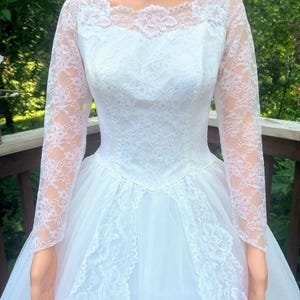 May include: A white wedding dress with long lace sleeves and a round neckline. The bodice and skirt are adorned with intricate lace details. The dress has a full, flowing skirt made of tulle.