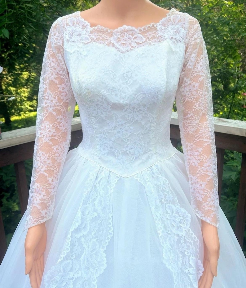 May include: A white wedding dress with long lace sleeves and a round neckline. The bodice and skirt are adorned with intricate lace details. The dress has a full, flowing skirt made of tulle.