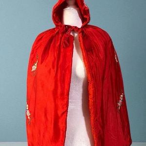 May include: A vibrant red cape with a hood, displayed on a mannequin. The cape features delicate floral embroidery in white, pink, and green. The hood is tied with a matching red ribbon.