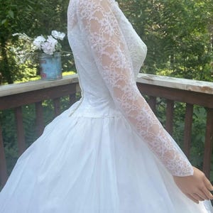 May include: White wedding dress with a lace bodice and long sleeves. The skirt is full and made of a sheer fabric. The dress is vintage and elegant, perfect for a special occasion.