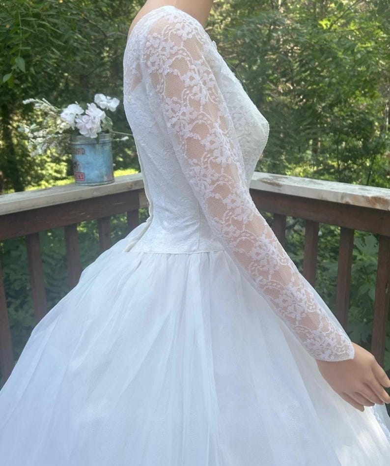 May include: White wedding dress with a lace bodice and long sleeves. The skirt is full and made of a sheer fabric. The dress is vintage and elegant, perfect for a special occasion.
