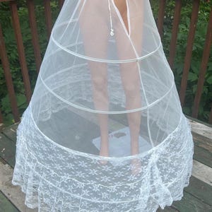 May include: A white crinoline petticoat with a lace hem. The petticoat has a tiered design with metal hoops for structure. The top is a sheer, lightweight fabric, and the bottom features a detailed floral lace trim.