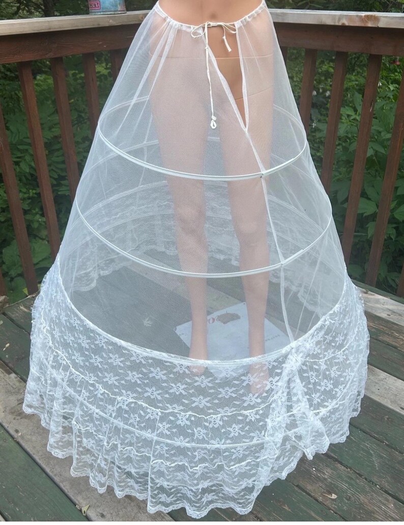 May include: A white crinoline petticoat with a lace hem. The petticoat has a tiered design with metal hoops for structure. The top is a sheer, lightweight fabric, and the bottom features a detailed floral lace trim.