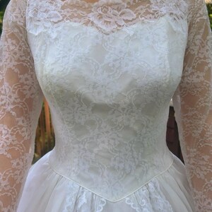 May include: A vintage white wedding dress featuring a lace bodice and long lace sleeves. The dress has a scoop neckline and a full skirt made of tulle. The intricate lace detailing adds a touch of elegance to this classic bridal gown.