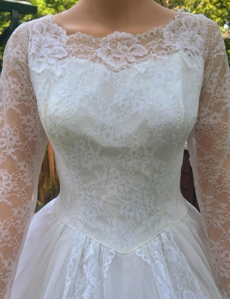 May include: A vintage white wedding dress featuring a lace bodice and long lace sleeves. The dress has a scoop neckline and a full skirt made of tulle. The intricate lace detailing adds a touch of elegance to this classic bridal gown.