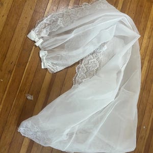 May include: A white bridal veil with delicate lace trim and small bows. The veil is made of a sheer, flowing fabric and is laid out on a wooden surface. The lace detailing adds a touch of elegance to the design.