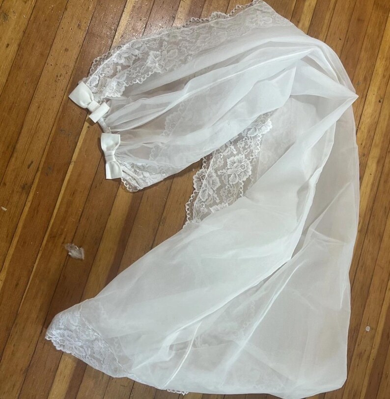 May include: A white bridal veil with delicate lace trim and small bows. The veil is made of a sheer, flowing fabric and is laid out on a wooden surface. The lace detailing adds a touch of elegance to the design.