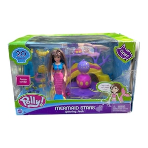 Op de afbeelding: Een Polly Pocket Mermaid Stars speelset in een doos. De set bevat een zeemeerminnenpop met een roze staart en blauwe top, en een draaiende zeehond. De doos heeft de tekst "20 stuks" en "Mermaid Stars".
