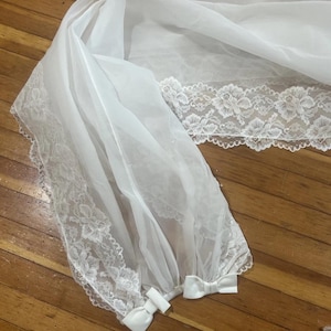 May include: A white, sheer veil with delicate floral lace trim along the edges. The veil is gathered at the bottom and tied with small white bows. The fabric appears lightweight and flowing, suitable for bridal wear or special occasions.