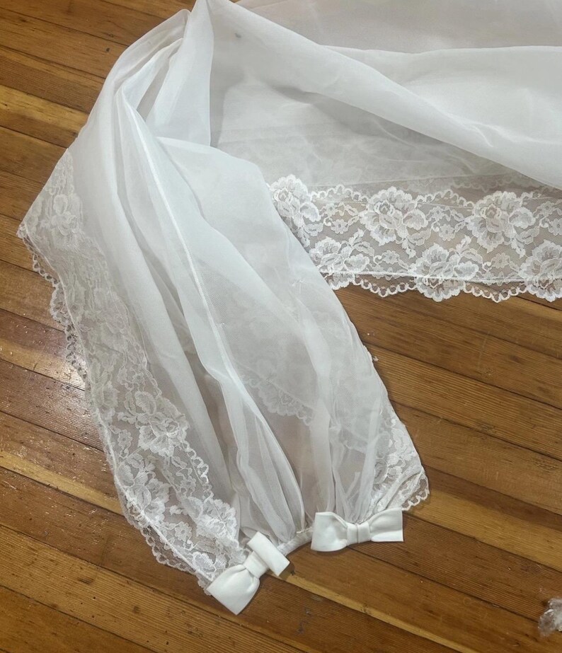 May include: A white, sheer veil with delicate floral lace trim along the edges. The veil is gathered at the bottom and tied with small white bows. The fabric appears lightweight and flowing, suitable for bridal wear or special occasions.