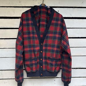 Op de afbeelding: Een vest met lange mouwen en een rood, marineblauw en groen ruitpatroon. Het kledingstuk heeft een effen marineblauwe V-hals, bijpassende geribbelde boorden en zoom, vijf bruine knopen aan de voorzijde en twee voorzakken.