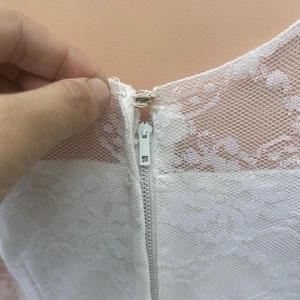 May include: Close-up of a white dress with a delicate lace overlay. The dress features a visible white zipper and a detailed lace neckline. The image highlights the texture and design of the fabric.