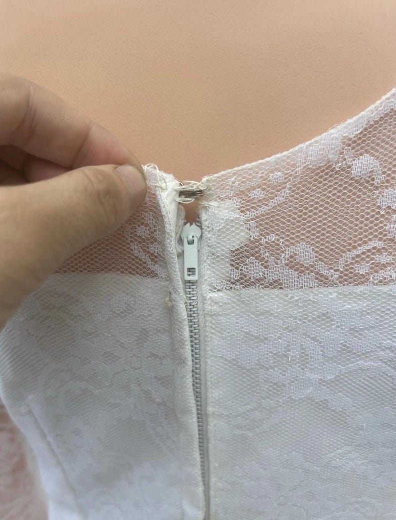 May include: Close-up of a white dress with a delicate lace overlay. The dress features a visible white zipper and a detailed lace neckline. The image highlights the texture and design of the fabric.