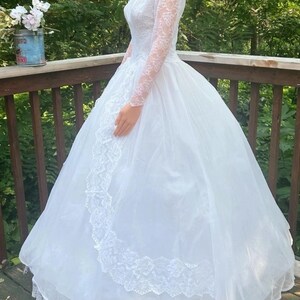 May include: A white wedding dress with a full skirt and long lace sleeves. The dress features a lace bodice and trim along the skirt. The dress is displayed outdoors on a wooden deck with a natural green background.