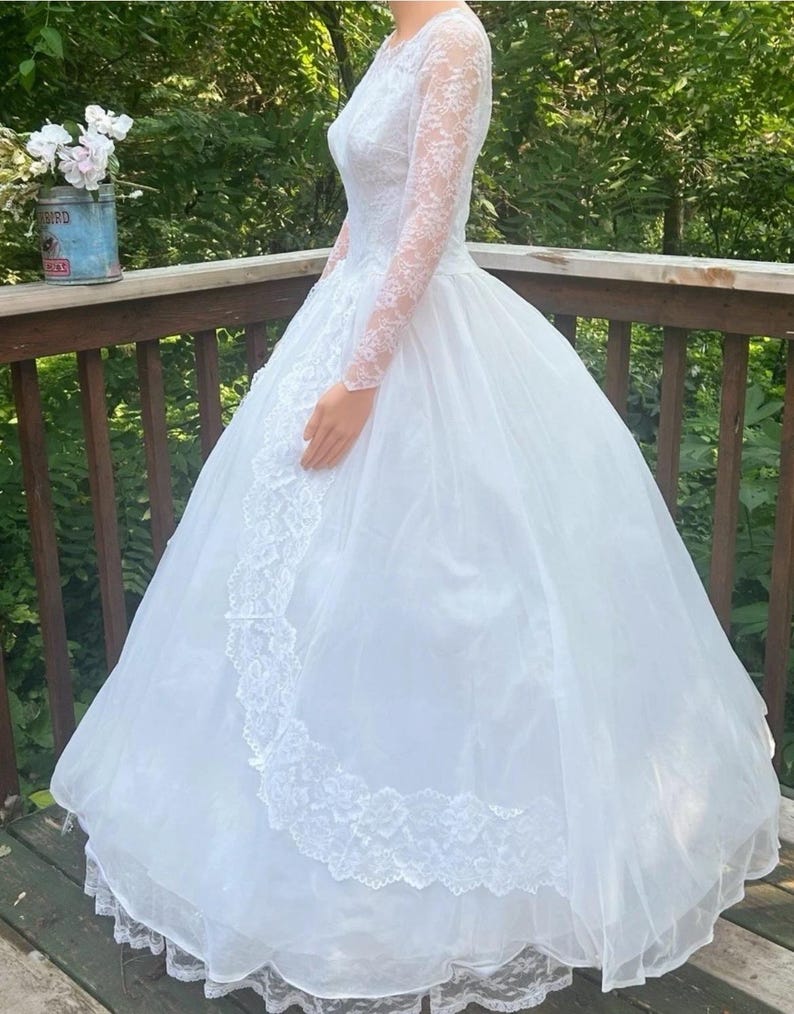 May include: A white wedding dress with a full skirt and long lace sleeves. The dress features a lace bodice and trim along the skirt. The dress is displayed outdoors on a wooden deck with a natural green background.