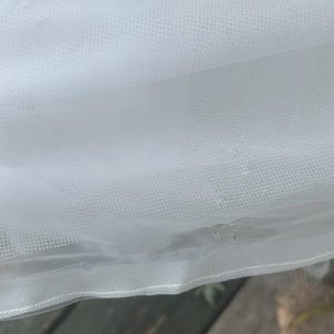 May include: Close-up of white tulle fabric. The fabric has a fine mesh texture and is slightly translucent. The edges are finished with a delicate, stitched hem. The fabric is draped, creating soft folds and shadows.