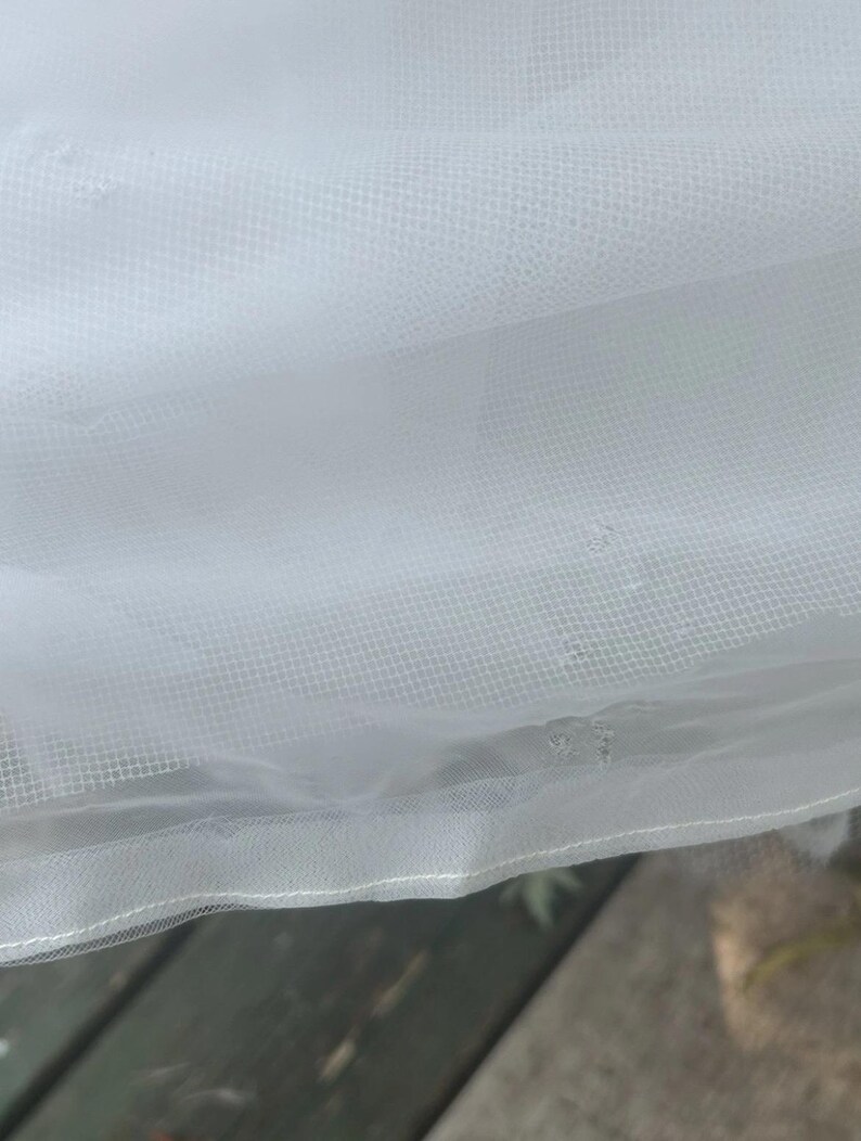 May include: Close-up of white tulle fabric. The fabric has a fine mesh texture and is slightly translucent. The edges are finished with a delicate, stitched hem. The fabric is draped, creating soft folds and shadows.