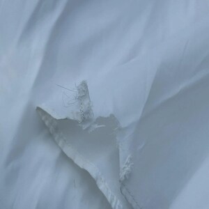 May include: Close-up of a white fabric with a torn edge. The fabric appears to be thin and slightly translucent, with visible threads and a frayed edge. The background is a soft, blurred white.