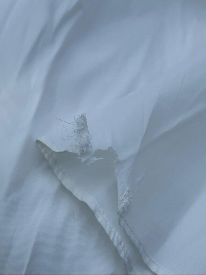May include: Close-up of a white fabric with a torn edge. The fabric appears to be thin and slightly translucent, with visible threads and a frayed edge. The background is a soft, blurred white.