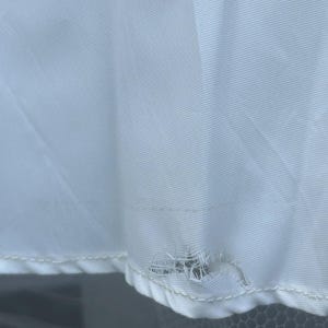 May include: Close-up of a white fabric with a tear near the bottom edge. The fabric has a subtle texture and a few small brown stains. The tear reveals the underlying structure of the fabric.