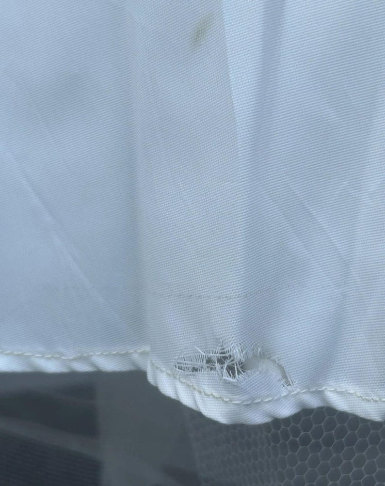 May include: Close-up of a white fabric with a tear near the bottom edge. The fabric has a subtle texture and a few small brown stains. The tear reveals the underlying structure of the fabric.