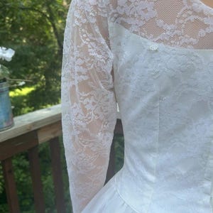 May include: White wedding dress with long lace sleeves. The bodice is fitted and the skirt flares out. The lace sleeves are sheer, and the dress appears to be made of a soft, flowing fabric. The dress is a classic style.