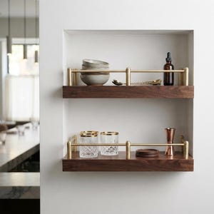 May include: Two horizontal wall-mounted shelves made of dark wood with brushed brass metal railings. The top shelf holds a stack of three ceramic bowls, a dark glass dropper bottle, and two gold spoons. The bottom shelf holds two crystal glasses, a copper jigger, and two dark wooden coasters.