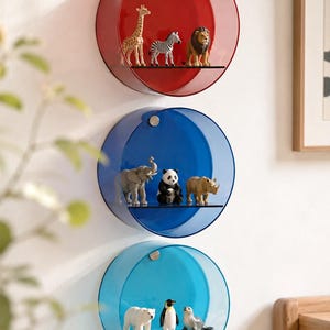 May include: Three circular wall-mounted shelves in red, blue, and light blue, each featuring a horizontal glass shelf. The red shelf holds giraffe, zebra, and lion figures. The blue shelf holds elephant, panda, and rhino figures. The light blue shelf holds polar bear, penguin, and seal figures.