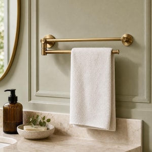 May include: A brushed brass finish metal towel rack with two horizontal bars mounted to the wall. One bar features a pivoting arm design, and a single white rectangular hand towel is draped over the lower bar, hanging vertically.