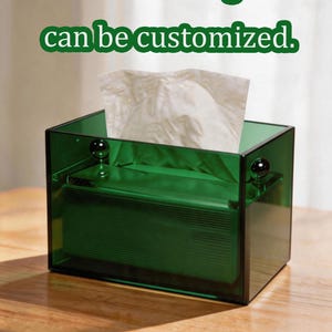 Op de afbeelding: Een rechthoekige donkergroene doorzichtige acryl tissuebox met een wit tissue dat aan de bovenkant uitsteekt. De doos heeft twee kleine zwarte bolvormige accenten op de bovenranden. De tekst "Text and logo can be customized." is zichtbaar.
