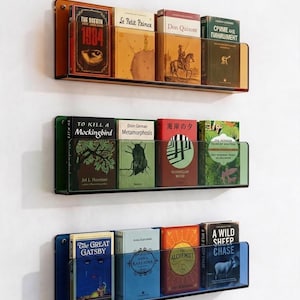 Könnte beinhalten: Drei horizontale Wandregale aus durchscheinendem Acryl in Bernstein, Grün und Blau. Jedes Regal verfügt über eine klare Frontplatte, um Bücher aufrecht zu halten. Die Regale sind vertikal angeordnet und mit verschiedenen Buchtiteln bestückt.